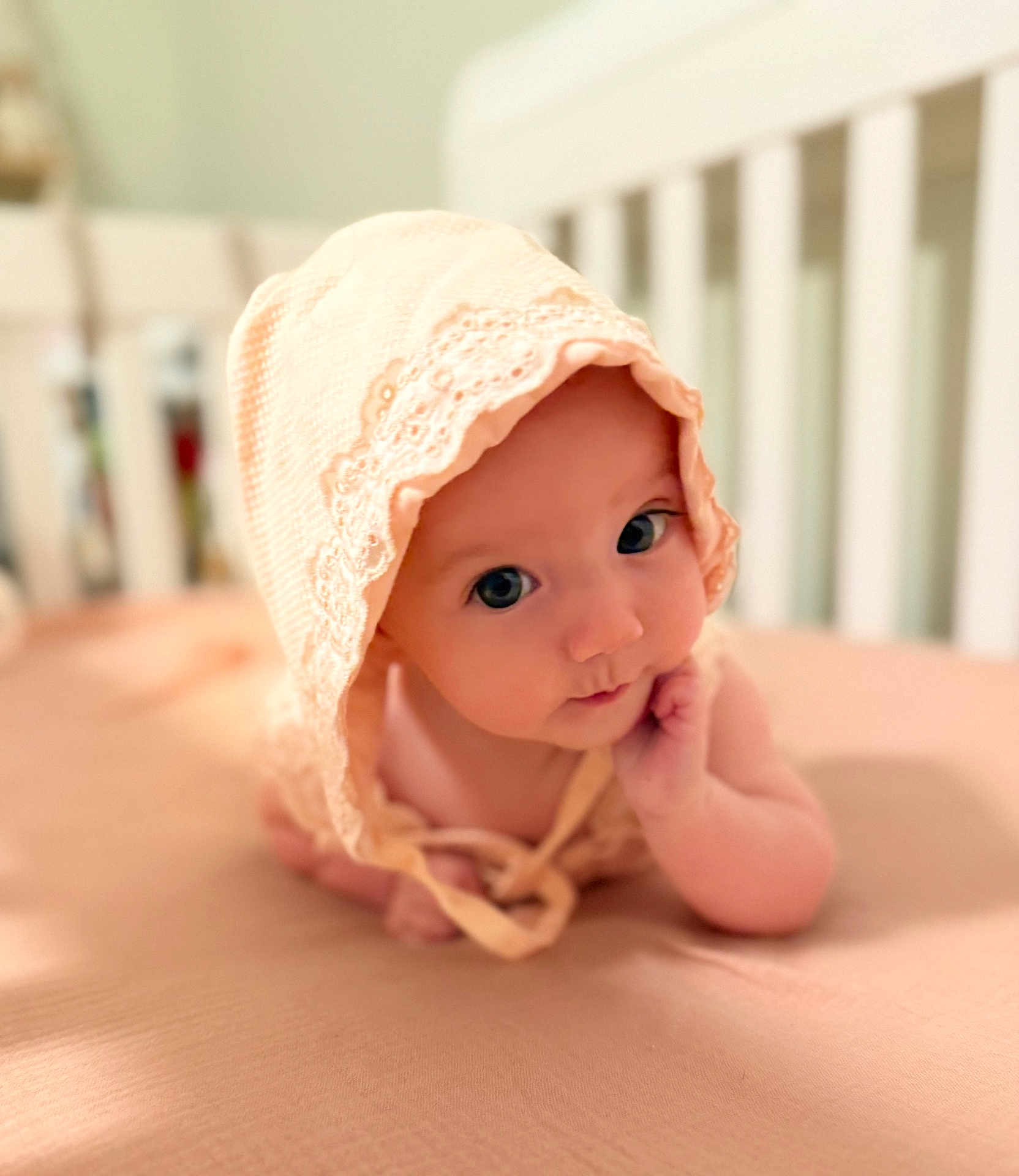Ailynn is registered to the contest to win money with this photo: baby, infant, bonnet, tummy_time, cute, child, portrait, soft_lighting, indoor, crib, bed, skin, hand, face, wide_eyes, peach, fabric, headwear, resting, curious