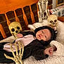 baby, baby_clothing, black_clothing, blanket, costume, cute, decor, face, halloween, indoor, infant, lying_down, pink_clothing, props, skeleton, skeleton_hand, skull, smiling, soft_texture, wooden_chair