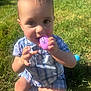 toddler, child, grass, outdoor, sunlight, toy, purple_egg, plaid_clothing, baby, sitting, chewing, hands, face, eyes, legs, shoes, greenery, daylight, playtime, infant