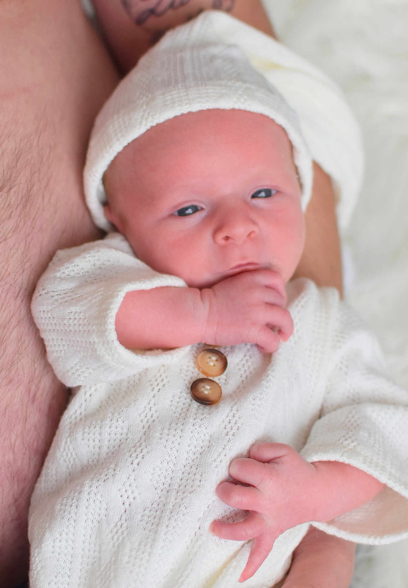 Liam a rejoint le concours — aidez-le/la à gagner de superbes lots ! newborn, baby, infant, white_clothing, knit_hat, knit_sweater, hand, fingers, resting, adult_arm, tattoo, skin, closeup, portrait, sleepy, cute, soft_light, indoor, cozy, peaceful