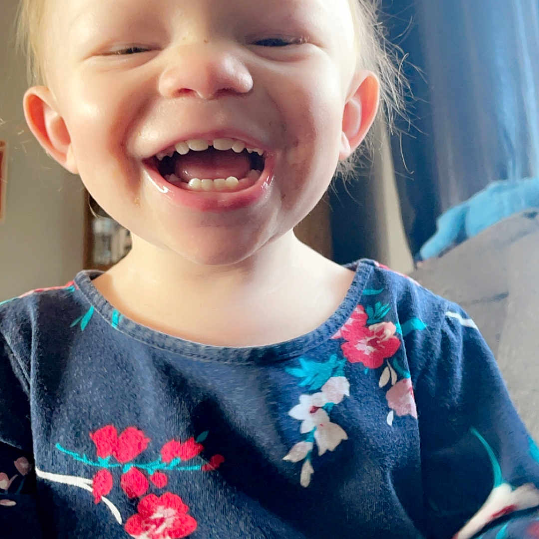 Delilah is registered to the contest to win money with this photo: accessories, baby, babylaughing, blonde, blouse, bodypart, clothing, dress, face, hair, happy, head, laughing, mouth, pajamas, person, photography, portrait, smile, teeth