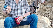 Hoss is registered to the contest to win money with this photo: beard, boots, cap, cup, dog, forest, gravel, harness, jeans, leash, outdoors, person, pitbull, plaid_shirt, relaxed_pose, rock, sitting, sunglasses, tattoo, watch