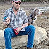 Hoss is registered to the contest to win money with this photo: beard, boots, cap, cup, dog, forest, gravel, harness, jeans, leash, outdoors, person, pitbull, plaid_shirt, relaxed_pose, rock, sitting, sunglasses, tattoo, watch