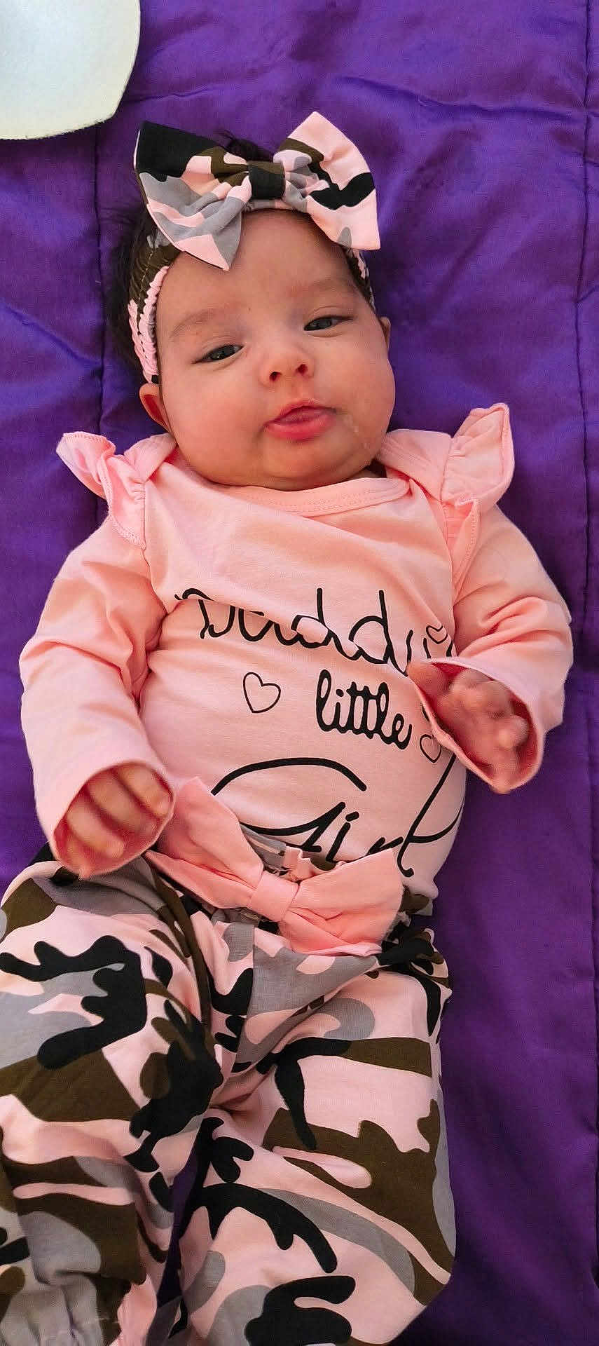 Kevani joined the competition — help win amazing prizes! baby, bow, camo_pattern, child, cute, drool, face, happy, headband, indoor, infant, long_sleeve, lying_down, pants, person, pink_clothing, portrait, purple_blanket, smile, toddler