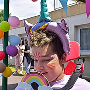 Leslie participe au concours pour gagner de l'argent avec cette photo : child, face_paint, balloon_hat, rainbow_prop, outdoor, festive, celebration, balloons, bunting, smiling, curly_hair, wheelchair, purple_shirt, sunlight, people, building, window, grass, daytime, fun