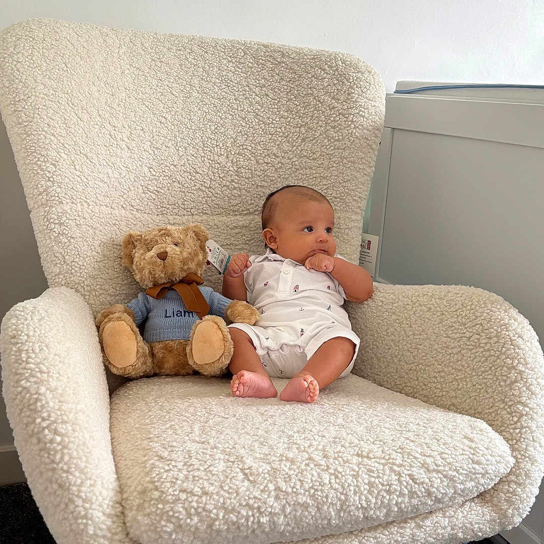 Liam is registered to the contest to win money with this photo: baby, carpet, child, clothing, cozy, cute, decor, foot, furniture, indoor, infant, person, plush_toy, rocking_chair, room, sitting, soft_texture, teddy_bear, toy, white_wall