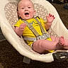 baby, child, yellow_onesie, baby_rocker, smiling, happy, indoor, carpet, patterned_cushion, sitting, straps, face, person, infant, toy, furniture, dog_cage, pet, home, vibration_control