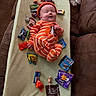 baby, child, candy, snickers, orange_pajamas, striped_clothing, blanket, couch, smiling, holding, wrapped_candy, lollipop, fun_size_candy, indoor, playful, cute, person, infant, lying_down, holiday