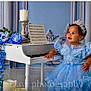 toddler, child, dress, blue_dress, tiara, piano, miniature_piano, flowers, blue_flowers, candles, music_sheet, decorative_wall, curly_hair, bracelet, portrait, indoor, smile, cute, celebration, elegant