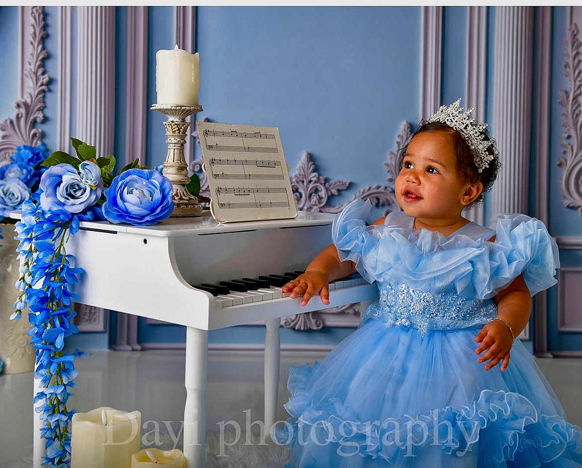 Ashley joined the competition — help win amazing prizes! toddler, child, dress, blue_dress, tiara, piano, miniature_piano, flowers, blue_flowers, candles, music_sheet, decorative_wall, curly_hair, bracelet, portrait, indoor, smile, cute, celebration, elegant