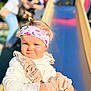 adult, baby, blurred_background, child, clothing, cute, daytime, fun, headband, outdoor, pattern, person, play, playground, ruffles, sitting, slide, smirk, sunlight, toddler