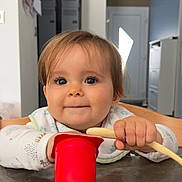 Léa a rejoint le concours — aidez-le/la à gagner de superbes lots ! baby, child, face, table, cup, spoon, bib, food, indoor, wood, hands, smile, eyes, clothing, furniture, kitchen, highchair, curly_hair, expression, cute