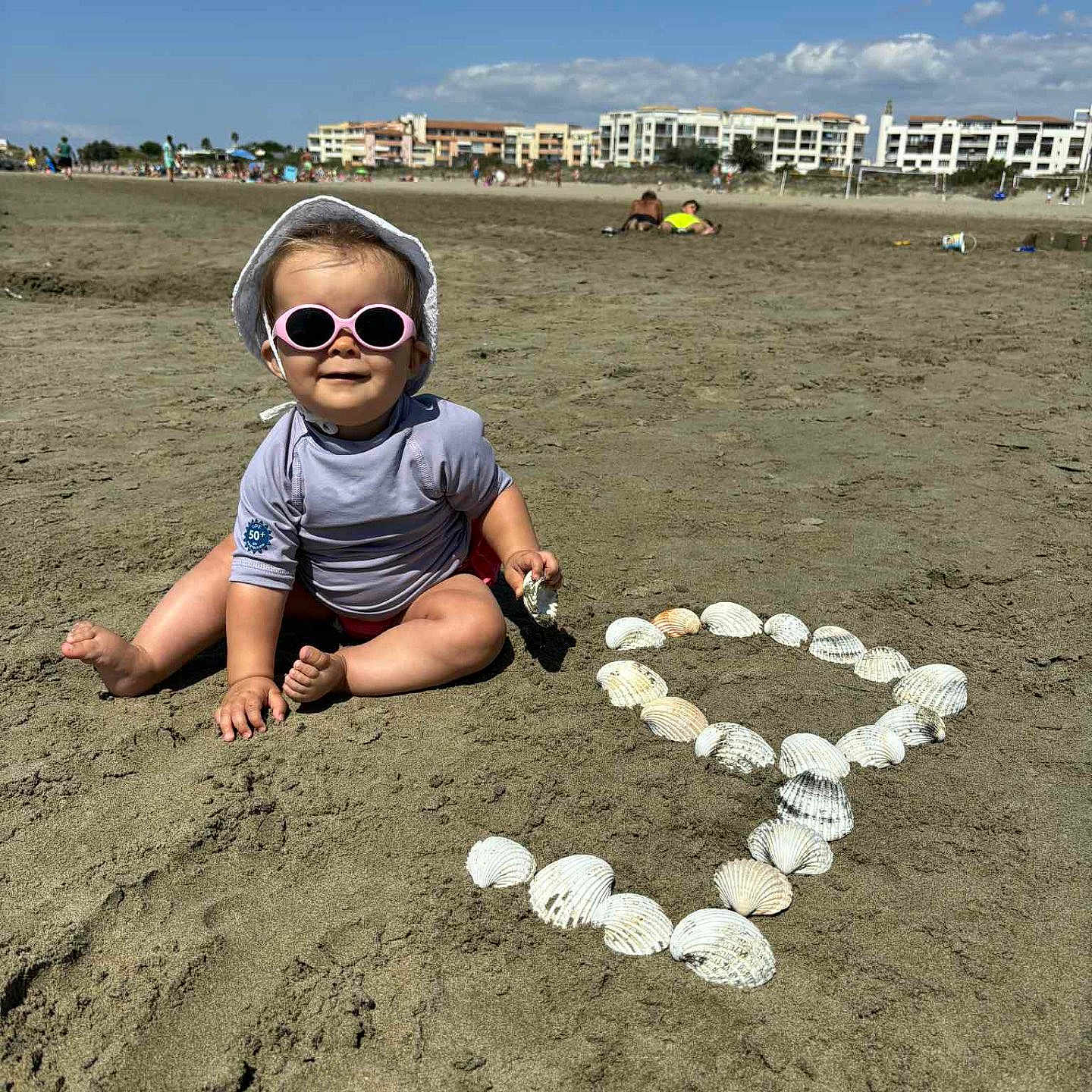 Léa participe au concours pour gagner de l'argent avec cette photo : beach, blue_sky, casual_clothing, child, coast, daytime, fun, happy, heart_shape, outdoor, playing, sand, seashells, smiling, summer, sun_hat, sunglasses, sunny, toddler, vacation