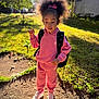 child, girl, outdoor, daylight, pink_clothing, hoodie, sweatpants, sneakers, backpack, curly_hair, hair_bow, standing, sunlight, shadow, grass, dirt_path, tree, residential_area, casual_wear, young_person