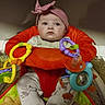 Bostyn is registered to the contest to win money with this photo: baby, child, infant, portrait, sitting, activity_center, jumper, pink_bow, toys, rattles, colorful, indoors, playtime, serious_expression, face, eyes, clothing, patterned_fabric, baby_gear, closeup