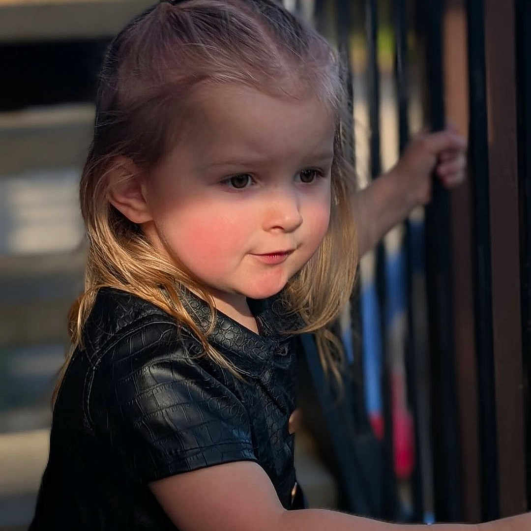Bostyn is registered to the contest to win money with this photo: child, toddler, girl, portrait, outdoors, staircase, railing, black_dress, black_bow, blonde_hair, rosy_cheeks, looking_sideways, standing, hand, face, hair, sunlight, portrait_photography, vertical_photo, safe