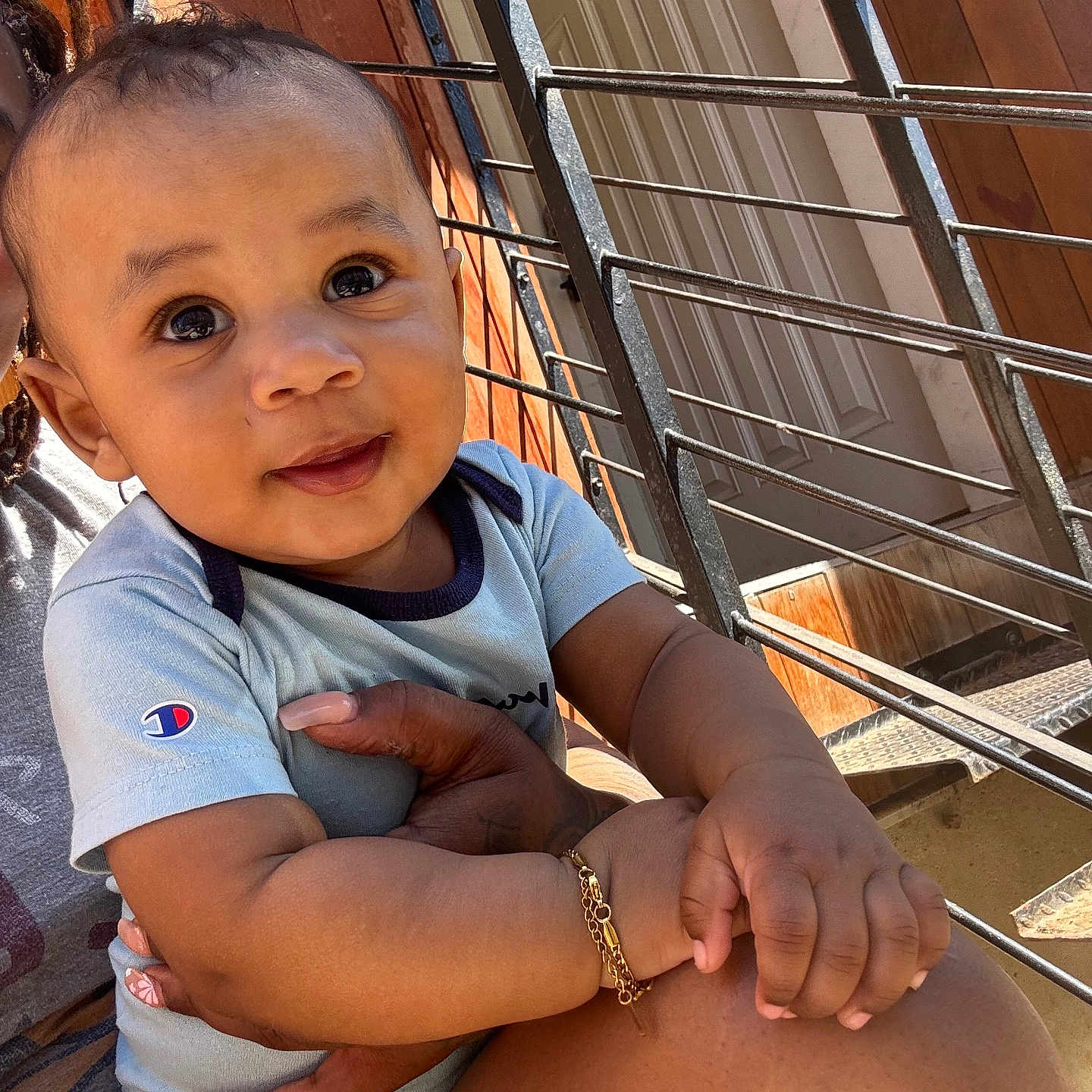 Malyk is registered to the contest to win money with this photo: baby, infant, child, person, smiling, blue_onesie, bracelet, hand, adult_hand, railing, wooden_wall, outdoor, porch, daylight, curious_eyes, closeup, portrait, holding, skin, face