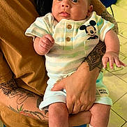 Liam joined the competition — help win amazing prizes! baby, infant, person, tattoo, clothing, mickey_mouse, hand, face, eyes, socks, footwear, adult, holding, indoor, floor, tile, curious, cute, portrait, casual