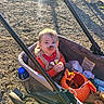 child, toddler, baby, stroller, outdoor, sunlight, face, portrait, serious_expression, lollipop, snack, red_clothing, vest, blond_hair, sitting, playground, dirt, ground, orange_bucket, water_bottle
