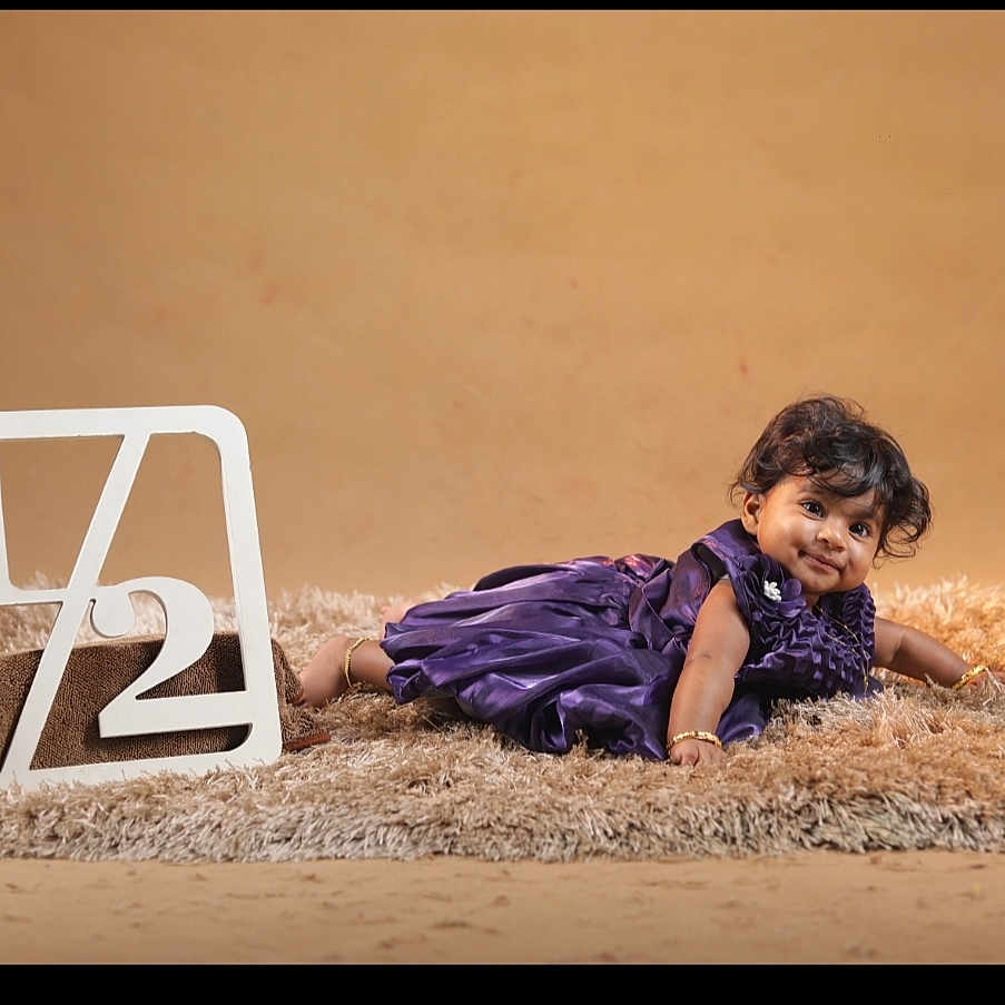 Paridhi joined the competition — help win amazing prizes! baby, beige_background, bracelet, celebration, child, curly_hair, cute, decorative_sign, face, floor, fluffy_rug, indoor, lying_down, milestone, person, portrait, purple_dress, smile, studio_photo, young_child
