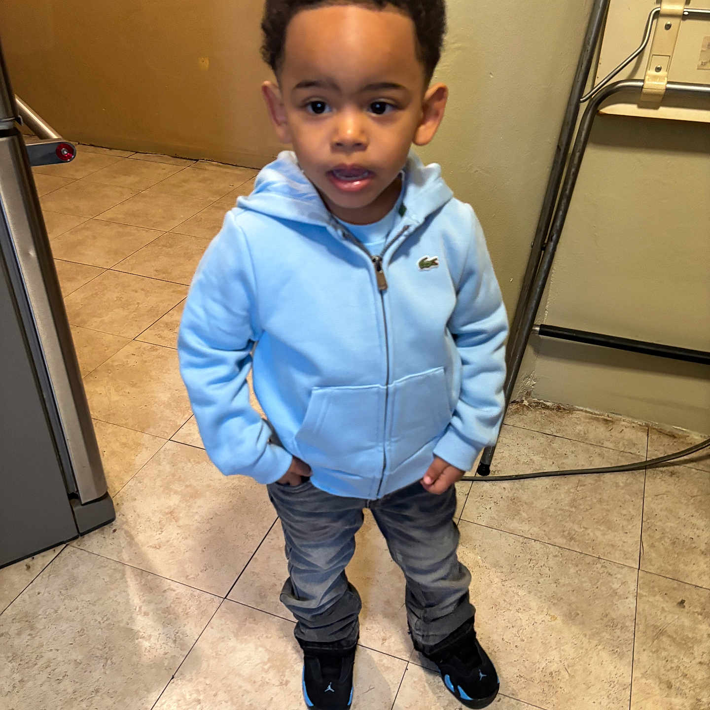 Karter is registered to the contest to win money with this photo: child, toddler, indoor, hoodie, blue_hoodie, jeans, black_shoes, curly_hair, standing, floor_tiles, neutral_wall, hand_in_pocket, casual_clothing, young_boy, expression, portrait, person, real_photo, home_interior, curious