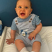 Noé participe au concours pour gagner de l'argent avec cette photo : baby, blue_wall, child, clothing, cushion, cute, donald_duck, face, footwear, hand, happy, indoor, infant, person, portrait, seated, smiling, socks, striped_overalls, white_shoes