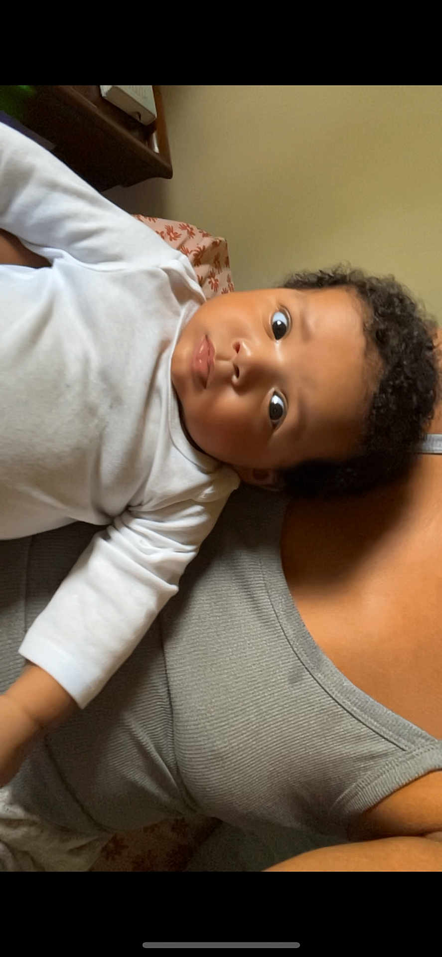 Messiah is registered to the contest to win money with this photo: baby, infant, face, person, child, curly_hair, white_clothing, gray_tank_top, cushion, patterned_cushion, indoor, closeup, portrait, skin, arm, head, expression, eyes, lying_down, comfort