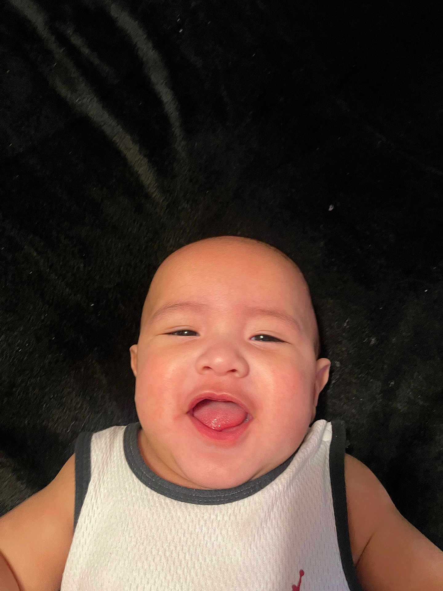Delijah joined the competition — help win amazing prizes! baby, infant, smiling, tongue_out, bald_head, white_tank_top, lying_down, black_blanket, face, chubby_cheeks, eyes, mouth, portrait, close_up, adorable, baby_clothing, playful, cute, infant_skin, cheeky