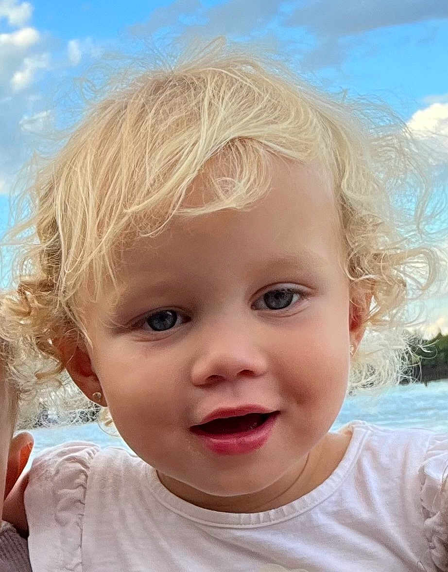 Tippa is registered to the contest to win money with this photo: toddler, child, blonde_hair, curly_hair, blue_eyes, smiling, face, outdoor, sky, clouds, water, white_shirt, earrings, portrait, young_child, cute, happy, person, natural_light, closeup