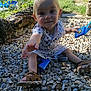 blue_toy, child, curious_expression, garden, greenery, nature, outdoor, patterned_clothing, pebbles, playful, reaching_hand, sandals, shadow, sitting, stone_wall, summer, sunlight, toddler, toy, young_child