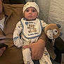 Lorenzo is registered to the contest to win money with this photo: baby, infant, pajamas, hat, bib, bear_pattern, plush_toy, adult_hand, chair, curtain, indoor, person, cute, wide_eyes, seated, clothing, child, parent, soft_lighting, portrait