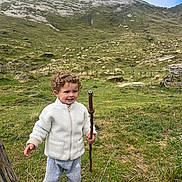 Elio a rejoint le concours — aidez-le/la à gagner de superbes lots ! child, toddler, curly_hair, white_jacket, gray_pants, black_shoes, walking_stick, mountain, grass, sheep, outdoor, nature, hill, fence, rocks, adventure, smile, person, daylight, scenery