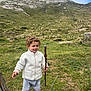 child, toddler, curly_hair, white_jacket, gray_pants, black_shoes, walking_stick, mountain, grass, sheep, outdoor, nature, hill, fence, rocks, adventure, smile, person, daylight, scenery