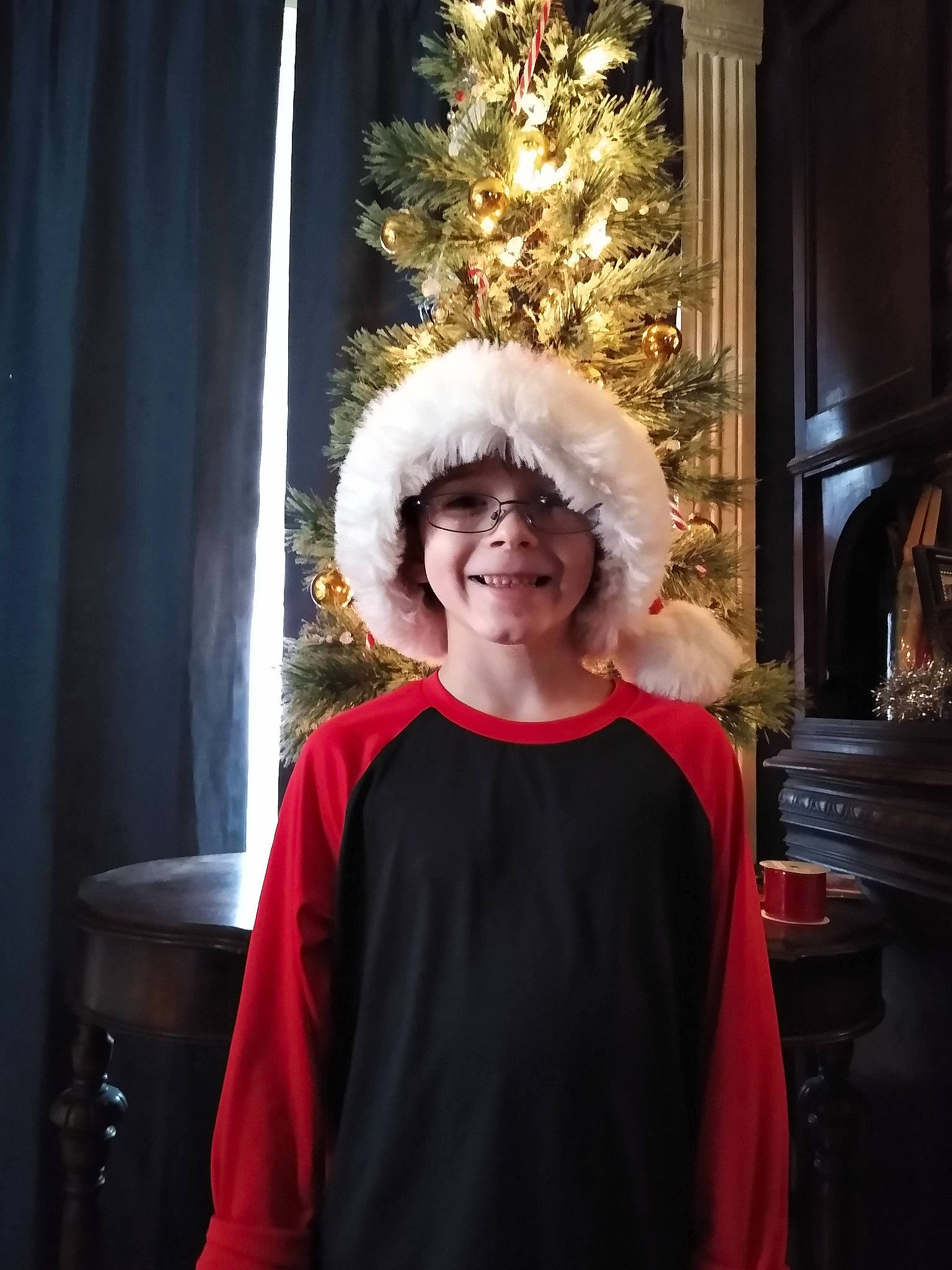 Logan is registered to the contest to win money with this photo: christmas_decoration, christmas_ornament, christmas_tree, curtain, event, fashion, fashion_design, formal_wear, fun, glasses, happy, hat, headwear, holiday, jewellery, joy, person, room, smile, standing