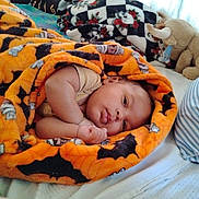 Xavier is registered to the contest to win money with this photo: baby, blanket, bat_pattern, orange, bed, pillow, striped_pillow, plush_toy, bull, infant, wrapped, cozy, sleeping_area, cute, soft_texture, child, lying_down, indoors, comfort, resting
