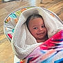 baby, blanket, colorful, smile, infant, face, fleece, wooden_floor, baby_seat, happy, indoors, person, wrapped, cozy, pet_legs, animal, flooring, child, cute, soft