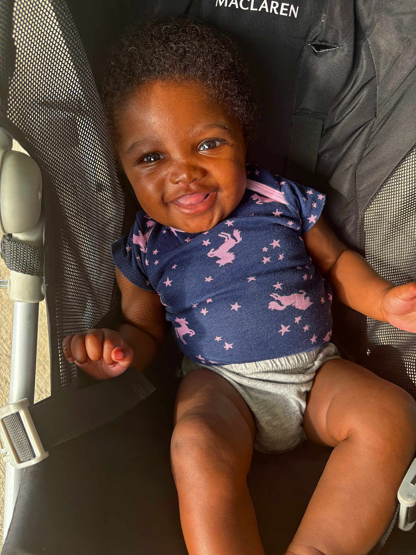 Eloni is registered to the contest to win money with this photo: baby, infant, child, smiling, happy, stroller, maclaren, blue_shirt, unicorn_pattern, shorts, sunlight, indoors, cute, cheerful, portrait, baby_clothing, legs, hands, seatbelt, mesh_fabric