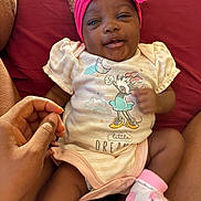 Eloni is registered to the contest to win money with this photo: baby, infant, smiling, headband, pink_bow, onesie, cartoon_print, hand, legs, socks, pink_socks, red_background, person, cute, closeup, indoors, child, happy, skin, toddler