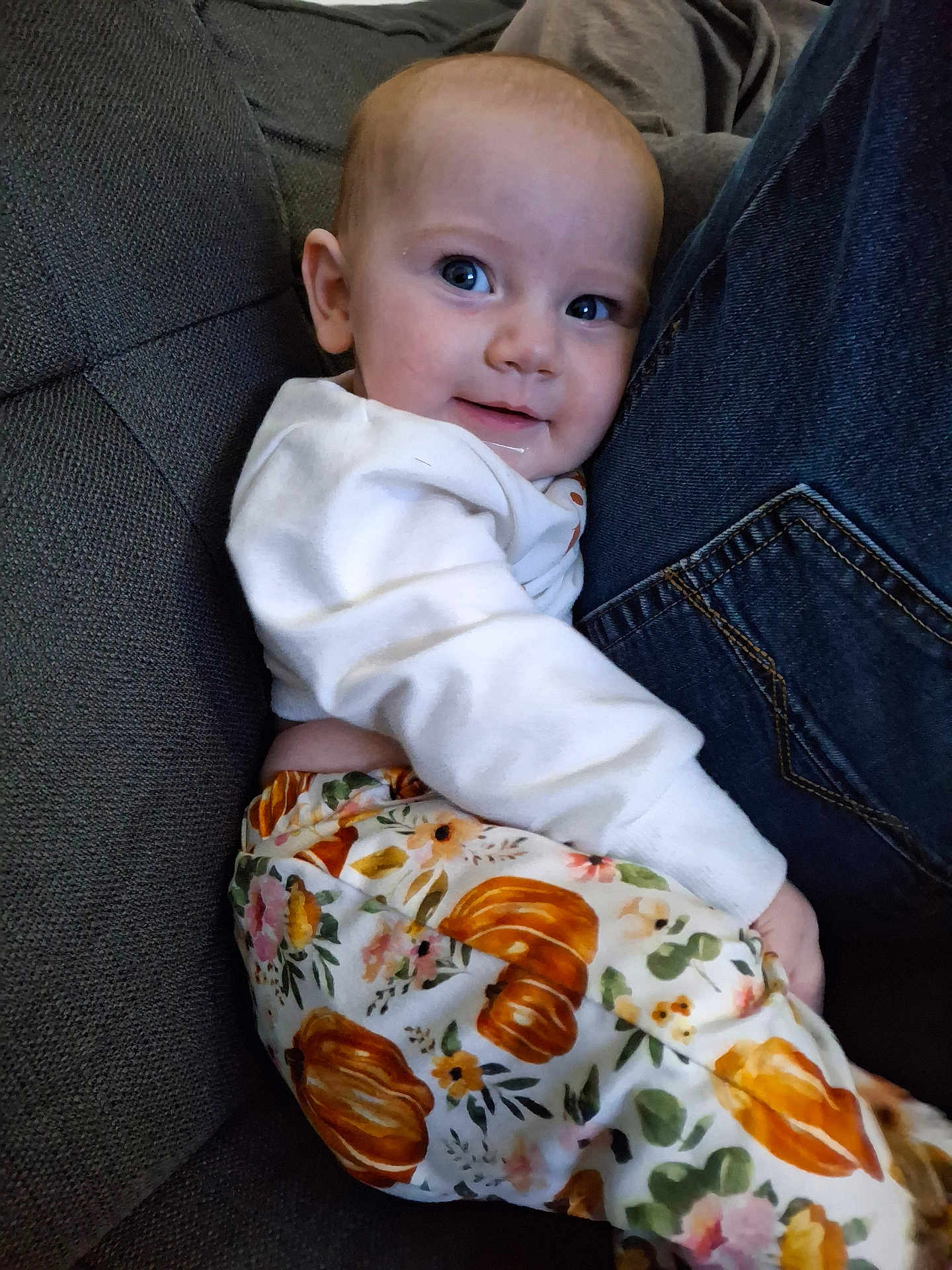 Anya is registered to the contest to win money with this photo: baby, child, person, jeans, couch, clothing, white_top, patterned_pants, pumpkin_pattern, floral_pattern, face, smile, indoor, seated, cozy, snuggling, cute, comfort, portrait, casual