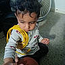 toddler, child, curly_hair, yellow_cable, hand_holding, carpet, fan, remote_control, indoor, sitting, long_sleeve_shirt, white_shirt, patterned_shirt, person, floor, technology, adult_hand, candid, casual, cute