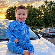 Henderson is registered to the contest to win money with this photo: child, toddler, baby, smiling, outdoor, sunset, car, vehicle, blue_outfit, jumpsuit, sitting, barefoot, portrait, parked_cars, trees, sky, clouds, golden_hour, parking_lot, happy