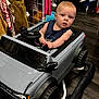 Jace joined the competition — help win amazing prizes! baby, child, toy_car, indoor, clothing, hanger, floor, steering_wheel, curious, blonde_hair, blue_eyes, sleeveless_shirt, plastic, wheels, flooring, shopping_area, apparel, seat, small_vehicle, playtime