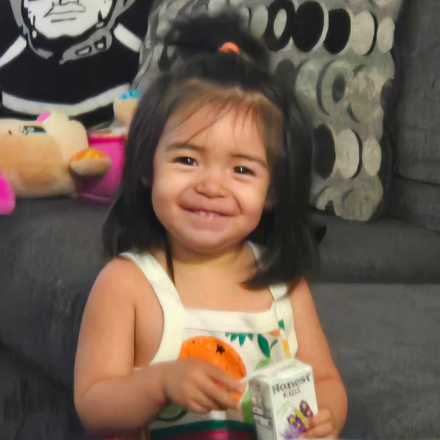 Roxanna is registered to the contest to win money with this photo: toddler, child, juice_box, smiling, sitting, couch, plush_toy, pillow, patterned_cushion, indoor, happy, person, black_and_white_art, hair_tie, tank_top, table, background_art, furniture, cute, portrait