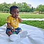toddler, child, outdoor, park, grass, blanket, yellow_shirt, denim_shorts, toy, sitting, greenery, nature, daylight, trees, person, footwear, casual_clothing, cute, curious, baby