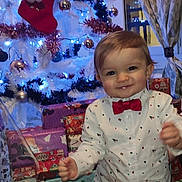 Ewenn a rejoint le concours — aidez-le/la à gagner de superbes lots ! toddler, child, christmas_tree, holiday, presents, bow_tie, festive_clothing, indoor, smiling, decorations, ornaments, lights, white_tree, curtains, window, wooden_floor, celebration, happy, party, cute