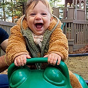 Sammy is registered to the contest to win money with this photo: toddler, child, smiling, outdoor, playground, play_equipment, green, frog_shaped_toy, jacket, happy, person, hands, wooden_structure, trees, nature, daylight, fun, supporting_adult, playtime, excited