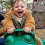 toddler, child, smiling, outdoor, playground, play_equipment, green, frog_shaped_toy, jacket, happy, person, hands, wooden_structure, trees, nature, daylight, fun, supporting_adult, playtime, excited