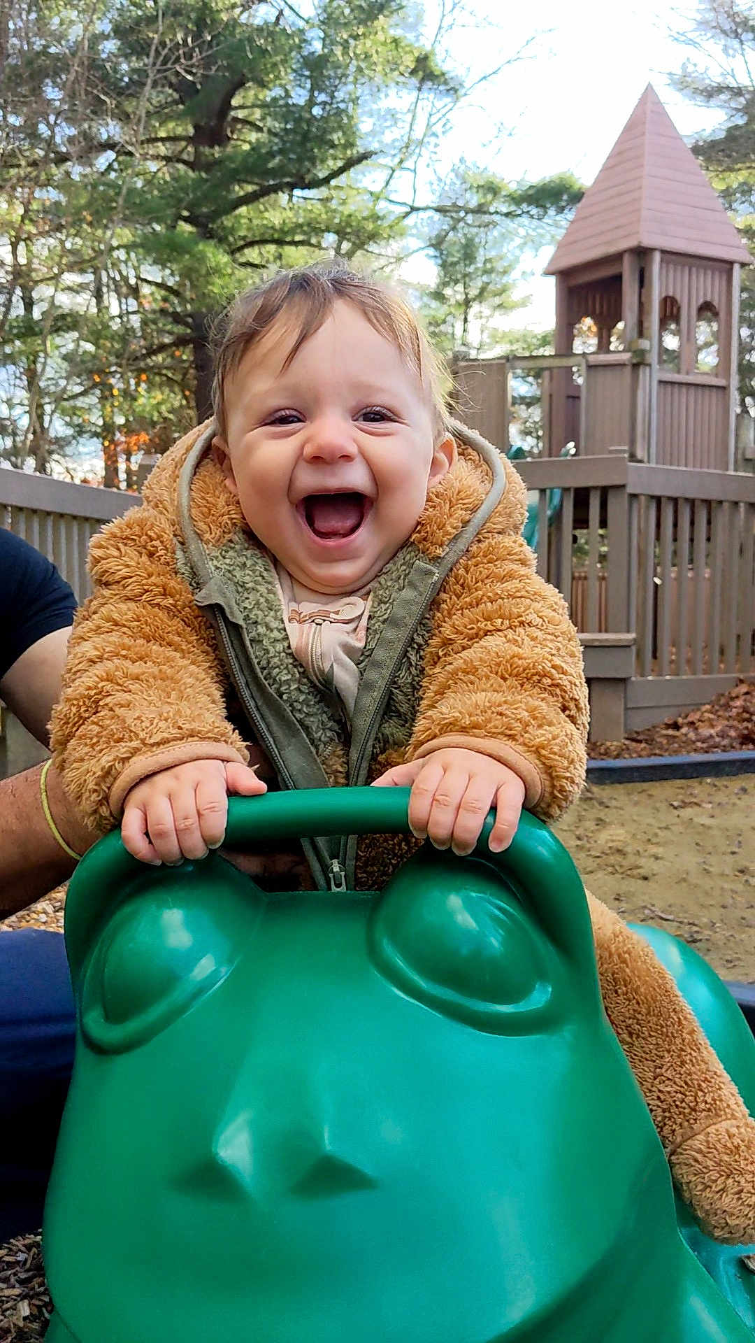 Sammy is registered to the contest to win money with this photo: toddler, child, smiling, outdoor, playground, play_equipment, green, frog_shaped_toy, jacket, happy, person, hands, wooden_structure, trees, nature, daylight, fun, supporting_adult, playtime, excited