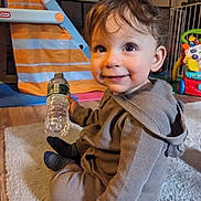 Sammy is registered to the contest to win money with this photo: child, toddler, infant, smile, water_bottle, toy, playroom, carpet, hoodie, curly_hair, big_eyes, sitting, indoors, floor, cute, happy, portrait, plastic_toy, activity_center, wooden_floor