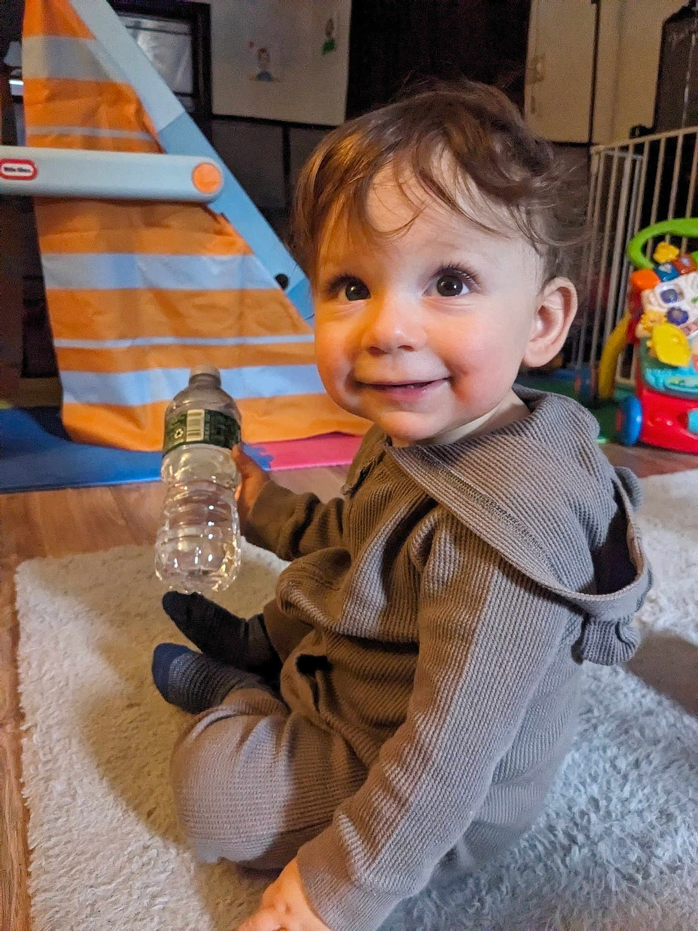 Sammy is registered to the contest to win money with this photo: child, toddler, infant, smile, water_bottle, toy, playroom, carpet, hoodie, curly_hair, big_eyes, sitting, indoors, floor, cute, happy, portrait, plastic_toy, activity_center, wooden_floor